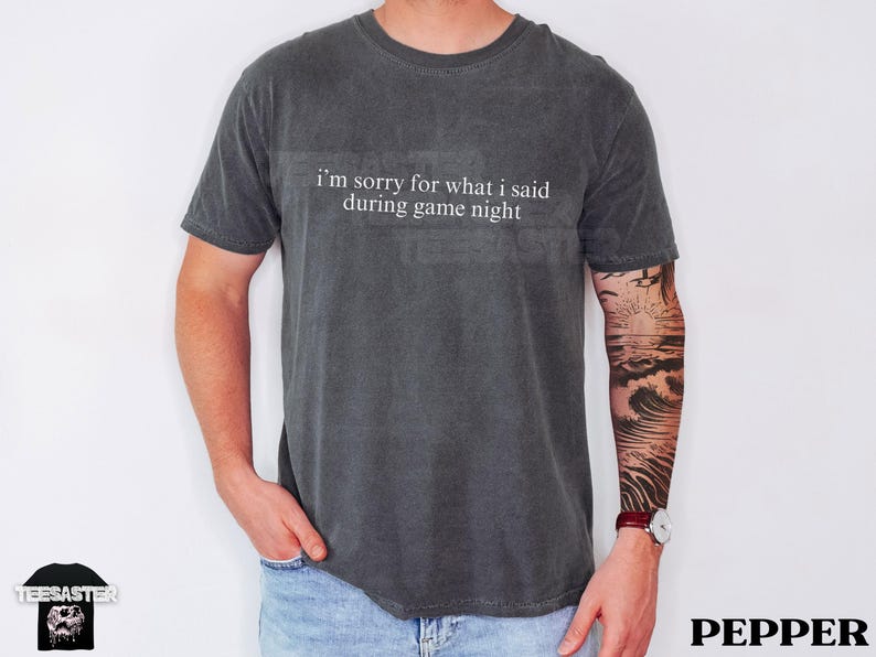 May include: Dark gray t-shirt with the text "I'm sorry for what I said during game night" in white. The shirt is a crew neck style and is worn by a person. The person is wearing blue jeans and has a tattoo on their arm.