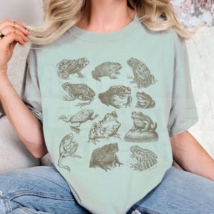 Pu&ograve; includere: T-shirt verde chiaro con illustrazioni dettagliate di rane in varie pose. Le rane sono raffigurate in uno stile botanico vintage, con una tavolozza di colori seppia sbiadita. La maglietta ha una vestibilit&agrave; rilassata.