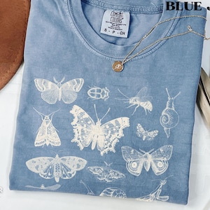 May include: Blue jean Comfort Colors t-shirt with a white insect and butterfly design. The shirt has a crew neck and short sleeves. A gold necklace with a pendant is draped across the shirt.