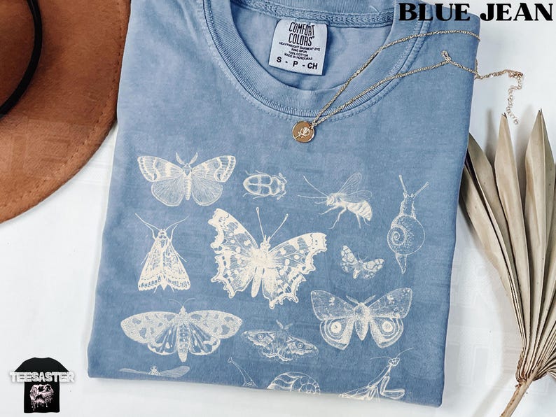 May include: Blue jean Comfort Colors t-shirt with a white insect and butterfly design. The shirt has a crew neck and short sleeves. A gold necklace with a pendant is draped across the shirt.