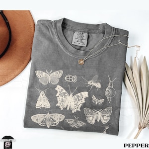 May include: A grey Comfort Colors t-shirt displaying insect illustrations, including butterflies, moths, and a beetle. The shirt has a crew neck and short sleeves. A silver necklace with a pendant rests on the shirt. The word "PEPPER" is at the bottom.