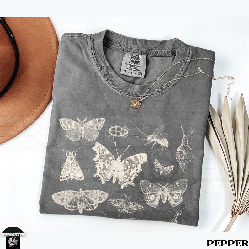 May include: A grey Comfort Colors t-shirt displaying insect illustrations, including butterflies, moths, and a beetle. The shirt has a crew neck and short sleeves. A silver necklace with a pendant rests on the shirt. The word "PEPPER" is at the bottom.