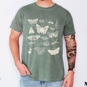 May include: Moss green t-shirt with a cream-coloured print of various insects, such as butterflies, moths, and beetles. The shirt has a crew neck and short sleeves. The word "TEESASTER" is printed on the bottom left. The word "MOSS" is printed on the bottom right.