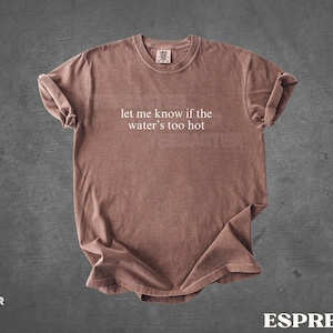 May include: Espresso-colored t-shirt with the text "let me know if the water's too hot" in white. The shirt is made of a soft material and has rolled-up sleeves. The shirt is displayed on a dark gray background.