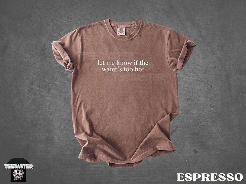 May include: Espresso-colored t-shirt with the text "let me know if the water's too hot" in white. The shirt is made of a soft material and has rolled-up sleeves. The shirt is displayed on a dark gray background.
