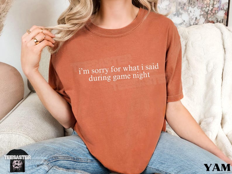 May include: A rust-colored t-shirt with the text "i'm sorry for what i said during game night" in white. The shirt is a short-sleeved, crew neck style. The image also shows a patch with a skull design.