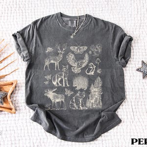 Pode incluir: T-shirt cinza escuro com uma ilustra&ccedil;&atilde;o creme de animais da floresta, incluindo um veado, uma coruja e uma raposa. O design tem um aspeto vintage. A palavra "PEPPER" est&aacute; impressa em branco. Gola redonda.