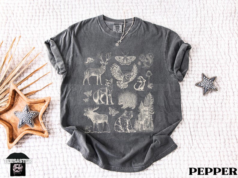 Pode incluir: T-shirt cinza escuro com uma ilustra&ccedil;&atilde;o creme de animais da floresta, incluindo um veado, uma coruja e uma raposa. O design tem um aspeto vintage. A palavra "PEPPER" est&aacute; impressa em branco. Gola redonda.