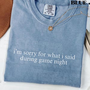May include: Blue jean-colored Comfort Colors t-shirt with the text "I'm sorry for what I said during game night" in white. A gold necklace with a pendant is on the shirt.