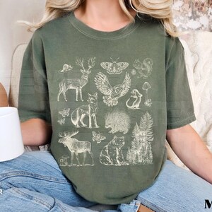 Pode incluir: Camiseta verde musgo com uma ilustra&ccedil;&atilde;o detalhada em creme de animais da floresta, incluindo um veado, uma coruja, uma raposa, um urso e um porco-espinho. A palavra "MOSS" est&aacute; impressa em preto no canto inferior direito.