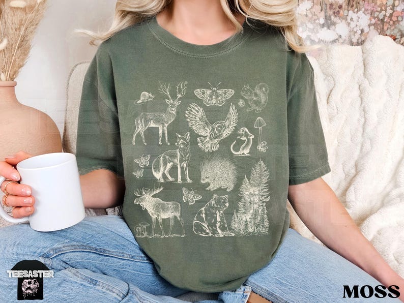 Pode incluir: Camiseta verde musgo com uma ilustra&ccedil;&atilde;o detalhada em creme de animais da floresta, incluindo um veado, uma coruja, uma raposa, um urso e um porco-espinho. A palavra "MOSS" est&aacute; impressa em preto no canto inferior direito.