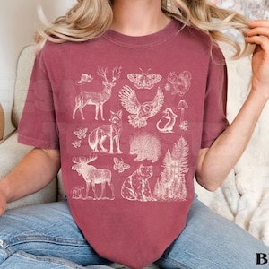 Pode incluir: Camiseta cor tijolo com uma ilustra&ccedil;&atilde;o branca de animais da floresta, incluindo um veado, uma coruja e uma raposa. A camiseta tem gola redonda e mangas curtas. A palavra "BRICK" est&aacute; impressa em branco no canto inferior direito.