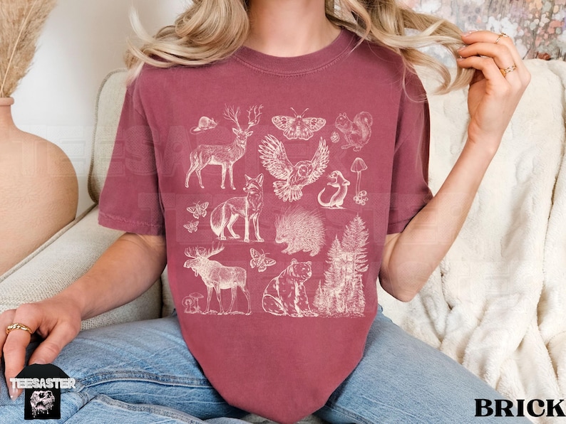 Pode incluir: Camiseta cor tijolo com uma ilustra&ccedil;&atilde;o branca de animais da floresta, incluindo um veado, uma coruja e uma raposa. A camiseta tem gola redonda e mangas curtas. A palavra "BRICK" est&aacute; impressa em branco no canto inferior direito.