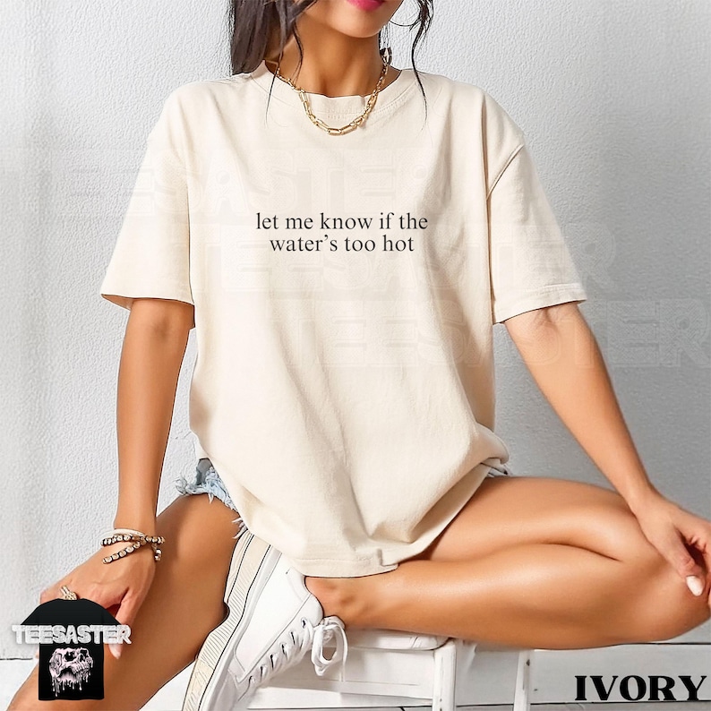 May include: Ivory-colored t-shirt with the text "let me know if the water's too hot" in black. The shirt is a relaxed fit. The image also shows a person wearing the shirt, sitting on a white stool.