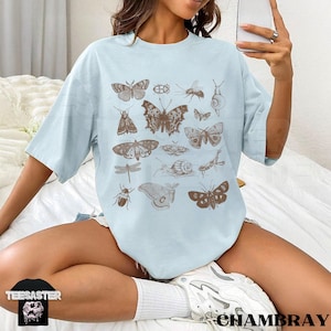 May include: A pale blue t-shirt displaying brown illustrations of various insects, such as butterflies, beetles, and a dragonfly. The shirt has short sleeves and a loose fit. The brand name "TEESASTER" is visible on a black tag.