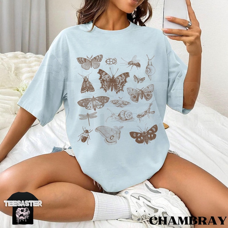 May include: A pale blue t-shirt displaying brown illustrations of various insects, such as butterflies, beetles, and a dragonfly. The shirt has short sleeves and a loose fit. The brand name "TEESASTER" is visible on a black tag.