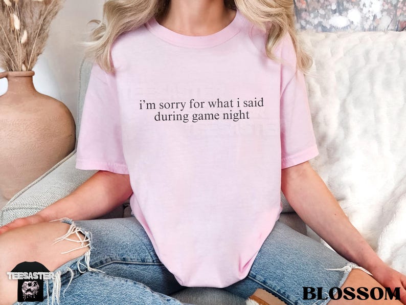 May include: A light pink t-shirt with the text "I'm sorry for what I said during game night" in a black, simple font. The shirt is worn with blue jeans. The word "BLOSSOM" is printed in the bottom right corner.
