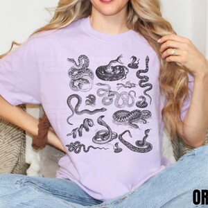 May include: A lilac t-shirt with a black and white illustration of different snakes. The design includes various snake species in detailed drawings. The word "ORCHID" is printed in black at the bottom right of the shirt.