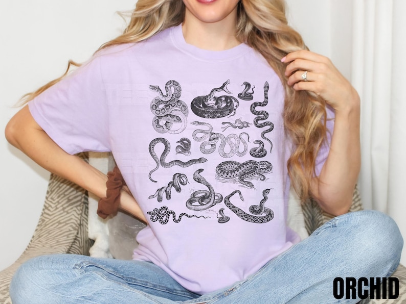 May include: A lilac t-shirt with a black and white illustration of different snakes. The design includes various snake species in detailed drawings. The word "ORCHID" is printed in black at the bottom right of the shirt.