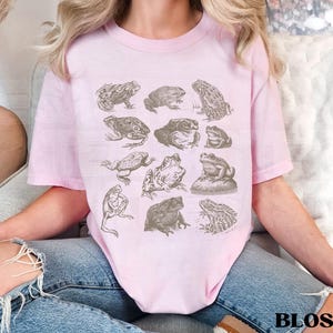 Pu&ograve; includere: Una maglietta rosa chiaro con illustrazioni dettagliate di rane in varie pose. La maglietta ha un girocollo e maniche corte. La parola "BLOSSOM" &egrave; stampata in nero in basso a destra.