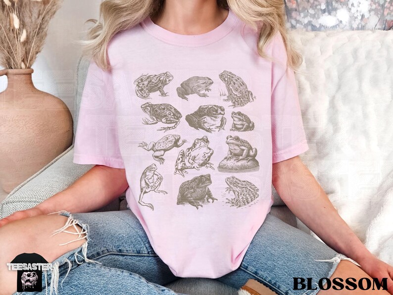 Pu&ograve; includere: Una maglietta rosa chiaro con illustrazioni dettagliate di rane in varie pose. La maglietta ha un girocollo e maniche corte. La parola "BLOSSOM" &egrave; stampata in nero in basso a destra.