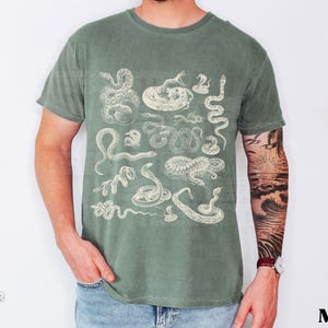 May include: Moss green t-shirt with a vintage-style illustration of various snakes. The design includes detailed drawings of different snake species. The word "TEESASTER" and a skull logo are on the bottom left.