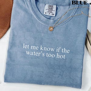 May include: Blue jean-colored t-shirt with the text "let me know if the water's too hot" in white. A gold necklace with a pendant rests on the shirt. The shirt is folded and has a Comfort Colors tag.