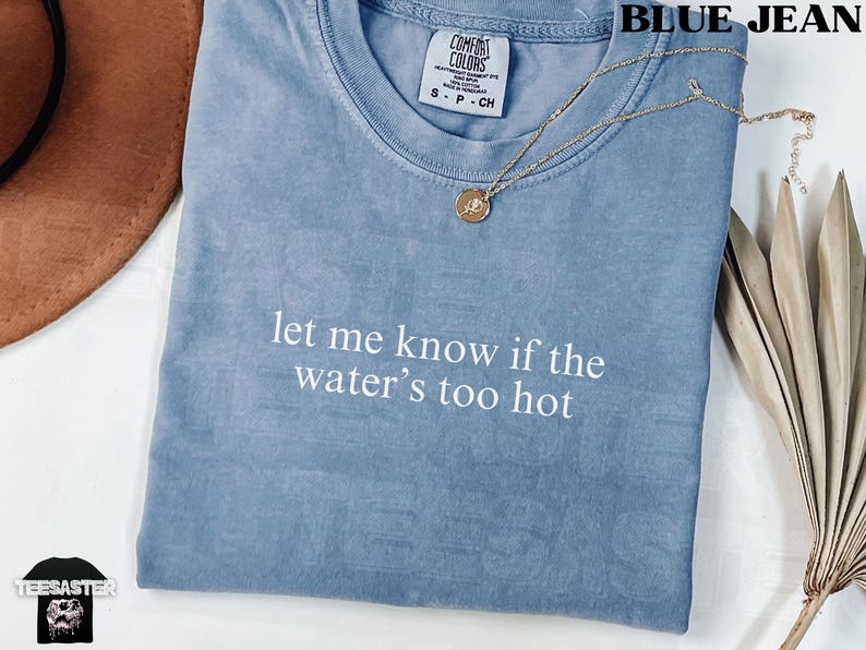 May include: Blue jean-colored t-shirt with the text "let me know if the water's too hot" in white. A gold necklace with a pendant rests on the shirt. The shirt is folded and has a Comfort Colors tag.