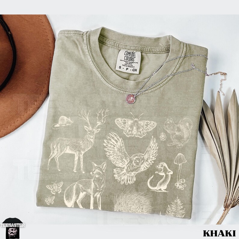 Pode incluir: Camiseta c&aacute;qui com gola redonda e mangas curtas. A camiseta apresenta um gr&aacute;fico branco de v&aacute;rios animais da floresta, incluindo um veado, uma coruja, uma raposa e um esquilo. Um colar de prata com um pingente est&aacute; sobre a camiseta.