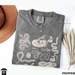 May include: A grey Comfort Colors t-shirt with various off-white snake illustrations. The shirt has a crew neck and short sleeves. A gold-coloured necklace with a pendant lies on the shirt. The word "PEPPER" is at the bottom.