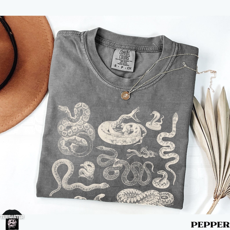 May include: A grey Comfort Colors t-shirt with various off-white snake illustrations. The shirt has a crew neck and short sleeves. A gold-coloured necklace with a pendant lies on the shirt. The word "PEPPER" is at the bottom.