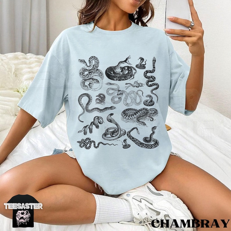 May include: Pale blue t-shirt with a print of numerous black and white snake illustrations. The shirt has a loose fit and short sleeves. The word "TEESASTER" is visible on a small black square at the bottom left.