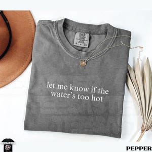 May include: A gray Comfort Colors t-shirt with the text "let me know if the water's too hot" in white. A silver necklace with a small pendant rests on the shirt. A brown hat and dried plant are in the background.
