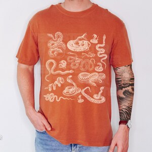 May include: Burnt orange t-shirt with white snake illustrations. The short-sleeved shirt has a crew neck. The shirt is worn by a person. The shirt is a solid colour with a vintage look.