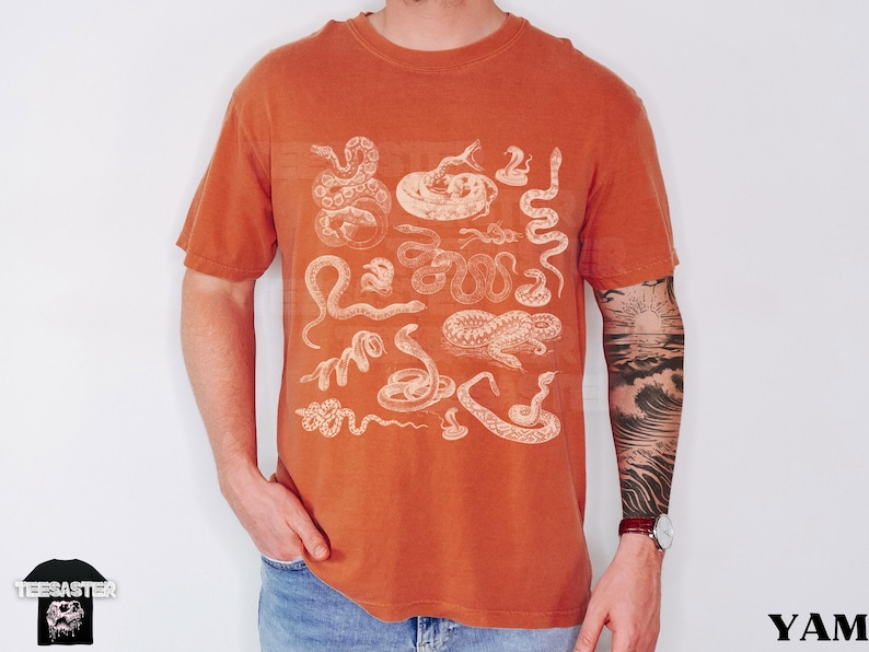 May include: Burnt orange t-shirt with white snake illustrations. The short-sleeved shirt has a crew neck. The shirt is worn by a person. The shirt is a solid colour with a vintage look.