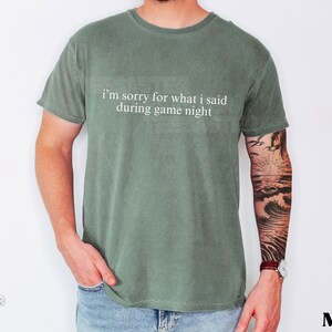 May include: Moss green t-shirt with the text "I'm sorry for what I said during game night" in white. The shirt is worn by a person wearing blue jeans. The word "Moss" is printed in the bottom right corner.