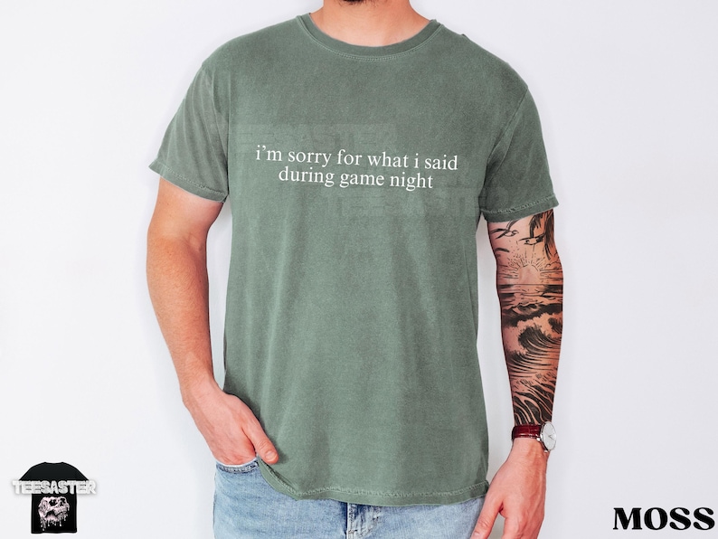 May include: Moss green t-shirt with the text "I'm sorry for what I said during game night" in white. The shirt is worn by a person wearing blue jeans. The word "Moss" is printed in the bottom right corner.