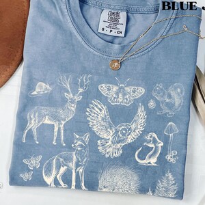 Pode incluir: Camiseta azul jeans Comfort Colors com um gr&aacute;fico branco com tema da natureza. O design inclui um veado, uma raposa, uma coruja, um esquilo e outros elementos da floresta. Um colar de ouro com um pingente repousa sobre a camisa.