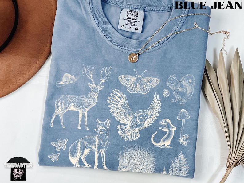 Pode incluir: Camiseta azul jeans Comfort Colors com um gr&aacute;fico branco com tema da natureza. O design inclui um veado, uma raposa, uma coruja, um esquilo e outros elementos da floresta. Um colar de ouro com um pingente repousa sobre a camisa.