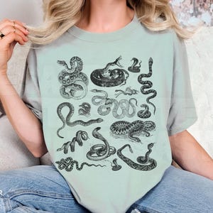 May include: A pale green t-shirt with a variety of black and white snake drawings. The top has a round neck and short sleeves. The brand logo is visible at the bottom left.