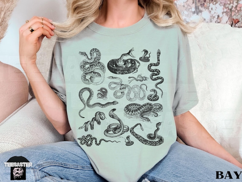 May include: A pale green t-shirt with a variety of black and white snake drawings. The top has a round neck and short sleeves. The brand logo is visible at the bottom left.