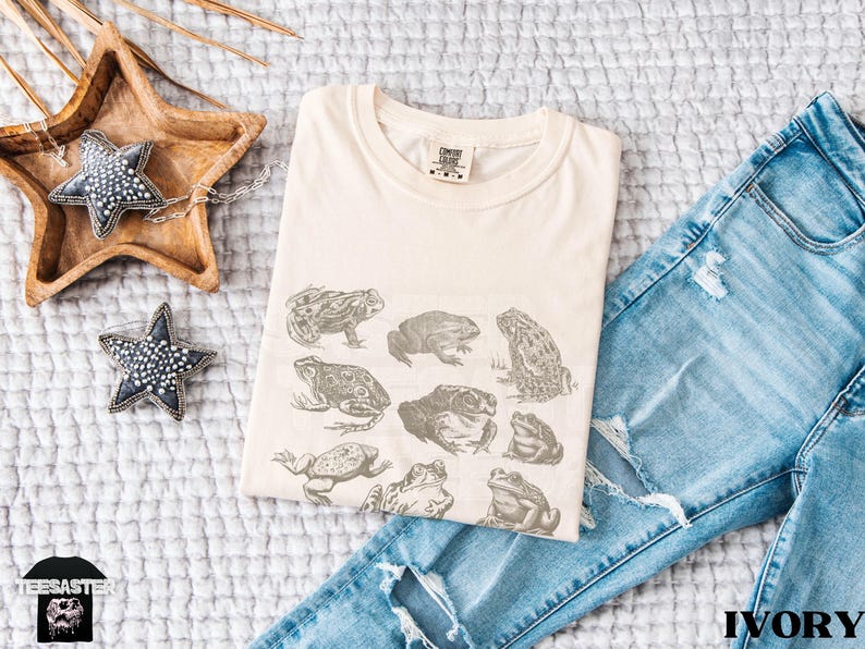 Pu&ograve; includere: T-shirt color avorio con un'illustrazione dettagliata di varie specie di rane. La maglietta &egrave; piegata accanto a un paio di jeans blu. Nell'immagine sono presenti anche oggetti decorativi a forma di stella e un vassoio di legno.