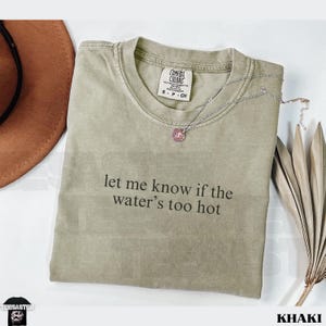 May include: Khaki-colored t-shirt with the text "let me know if the water's too hot." The shirt has a crew neck and short sleeves. A silver necklace with a pendant and a brown hat are next to the shirt.