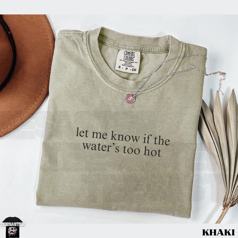 May include: Khaki-colored t-shirt with the text "let me know if the water's too hot." The shirt has a crew neck and short sleeves. A silver necklace with a pendant and a brown hat are next to the shirt.