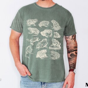 Pu&ograve; includere: T-shirt verde muschio con illustrazioni di rane in stile vintage. La maglietta ha un girocollo e maniche corte. La parola "Moss" &egrave; stampata in grassetto in basso a destra.