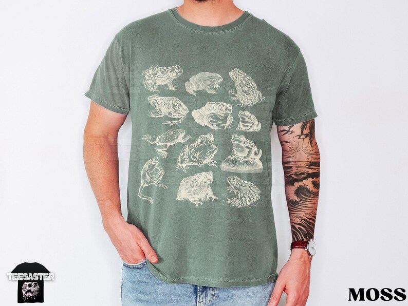 Pu&ograve; includere: T-shirt verde muschio con illustrazioni di rane in stile vintage. La maglietta ha un girocollo e maniche corte. La parola "Moss" &egrave; stampata in grassetto in basso a destra.