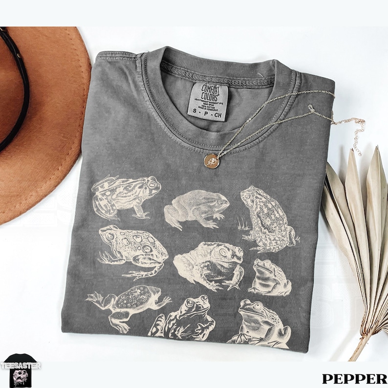 Pu&ograve; includere: Una maglietta grigia Comfort Colors con illustrazioni di rane bianche. La maglietta ha un girocollo e maniche corte. Una collana d'argento con un piccolo ciondolo &egrave; sulla maglietta. La parola "PEPPER" &egrave; nell'angolo in basso a destra.