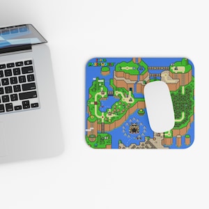 May include: A rectangular mousepad featuring a colorful map design from the Super Mario World video game. The mousepad has a blue background with green, brown, and yellow elements. A white computer mouse rests on the pad.