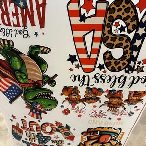 May include: A white surface covered in patriotic-themed graphics. The design includes the word "AMERICA" in red, a green dinosaur wearing a cape and an American flag, and the phrase "God Bless The USA". Other graphics include stars, fireworks, and the words "Chill Out".