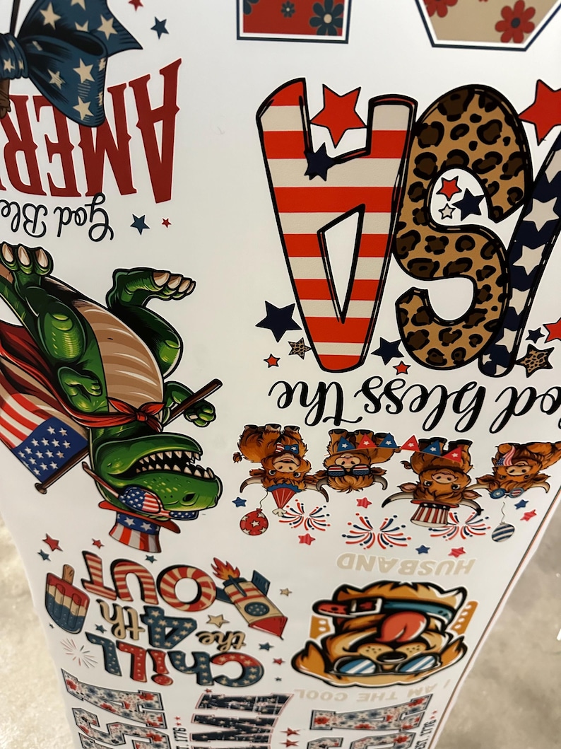 May include: A white surface covered in patriotic-themed graphics. The design includes the word "AMERICA" in red, a green dinosaur wearing a cape and an American flag, and the phrase "God Bless The USA". Other graphics include stars, fireworks, and the words "Chill Out".
